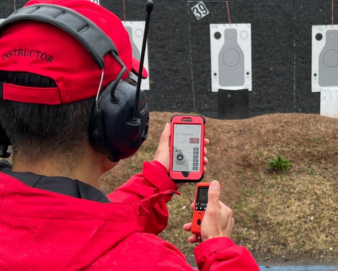 A range instructor holding a phone running JMAP, with a shot timer in the other hand