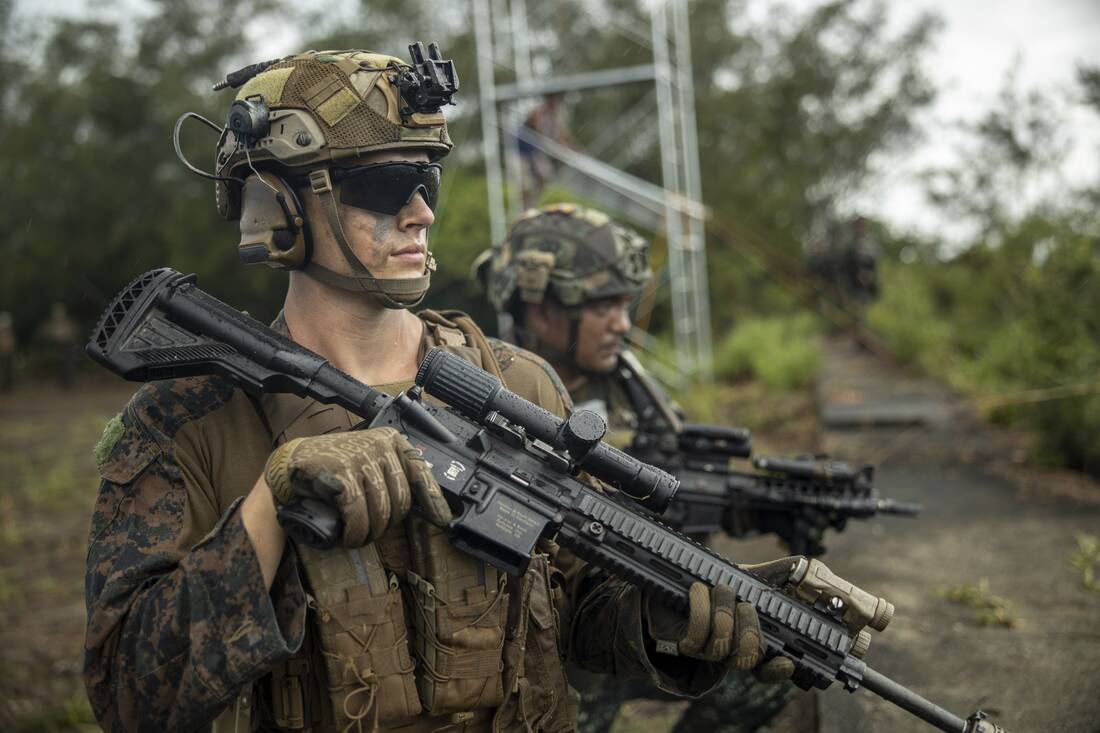Two Marines in helmets, eye protection and plate carriers holding rifles with optics