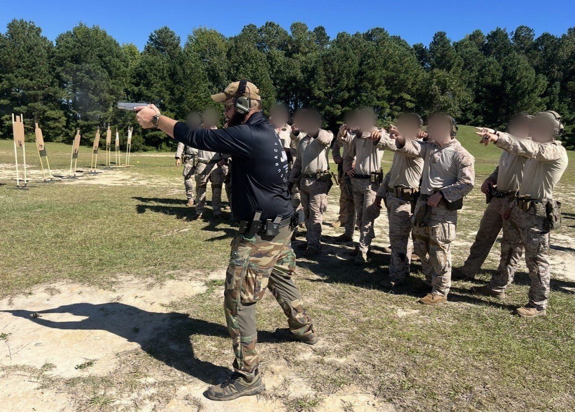 A Scoring Tech instructor firing a pistol on the line while a class of service members points at the targets; faces blurred