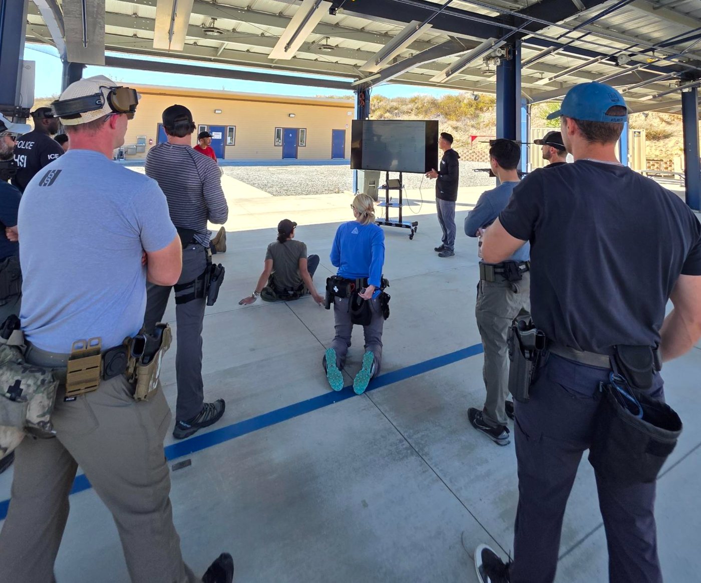 Law enforcement trainees gathered under a covered range as an instructor briefs assessment results on a large TV